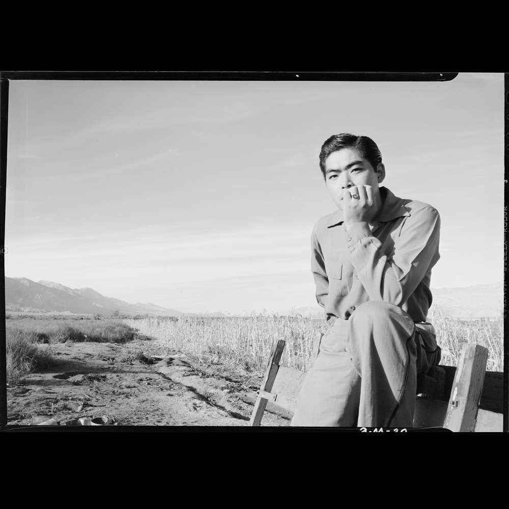 Tom Kobayashi, half-length portrait, seated in a field at the Manzanar Relocation Center, California, his elbow resting on his knee and his hand on his chin, facing front.