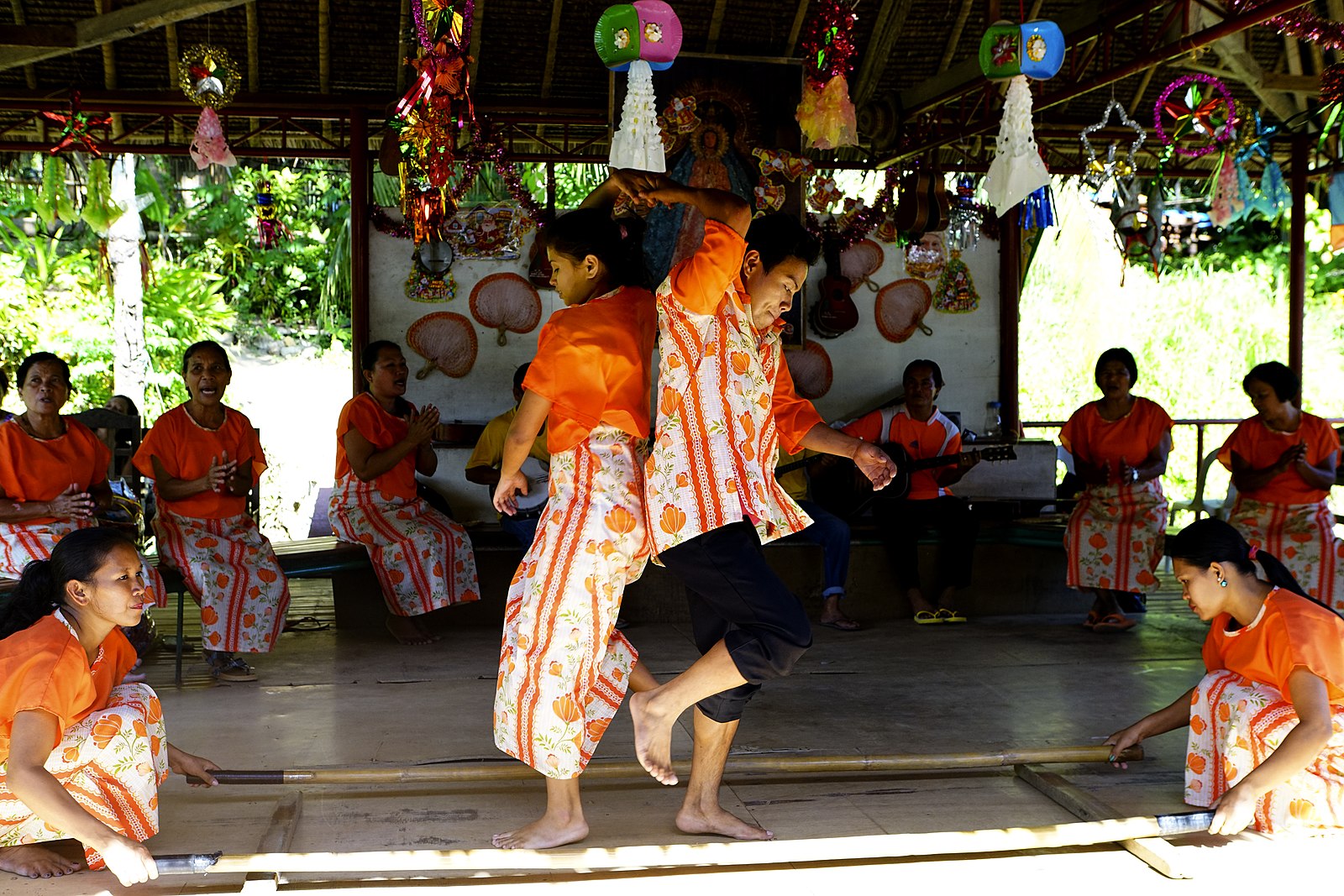Two individuals are holding hands and are facing opposite directions with their backs against one another in a popular folk dance from the Philippines called the Tinikling.
