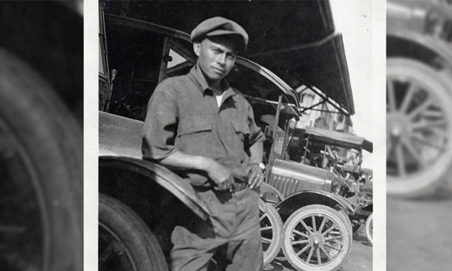 Black and white photograph of a Filipino mechanic in work clothes and a flat cap posing in a garage full of trucks.