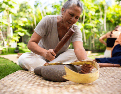 Artist Page Change seated on the ground making traditional Hawaiian barkcloth or &ldquo;kapa&rdquo; by softening mulberry bark on a stone with a wooden mallet.