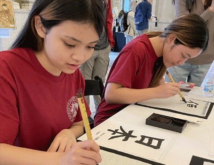 Two young students in red t-shirts practicing Japanese calligraphy.